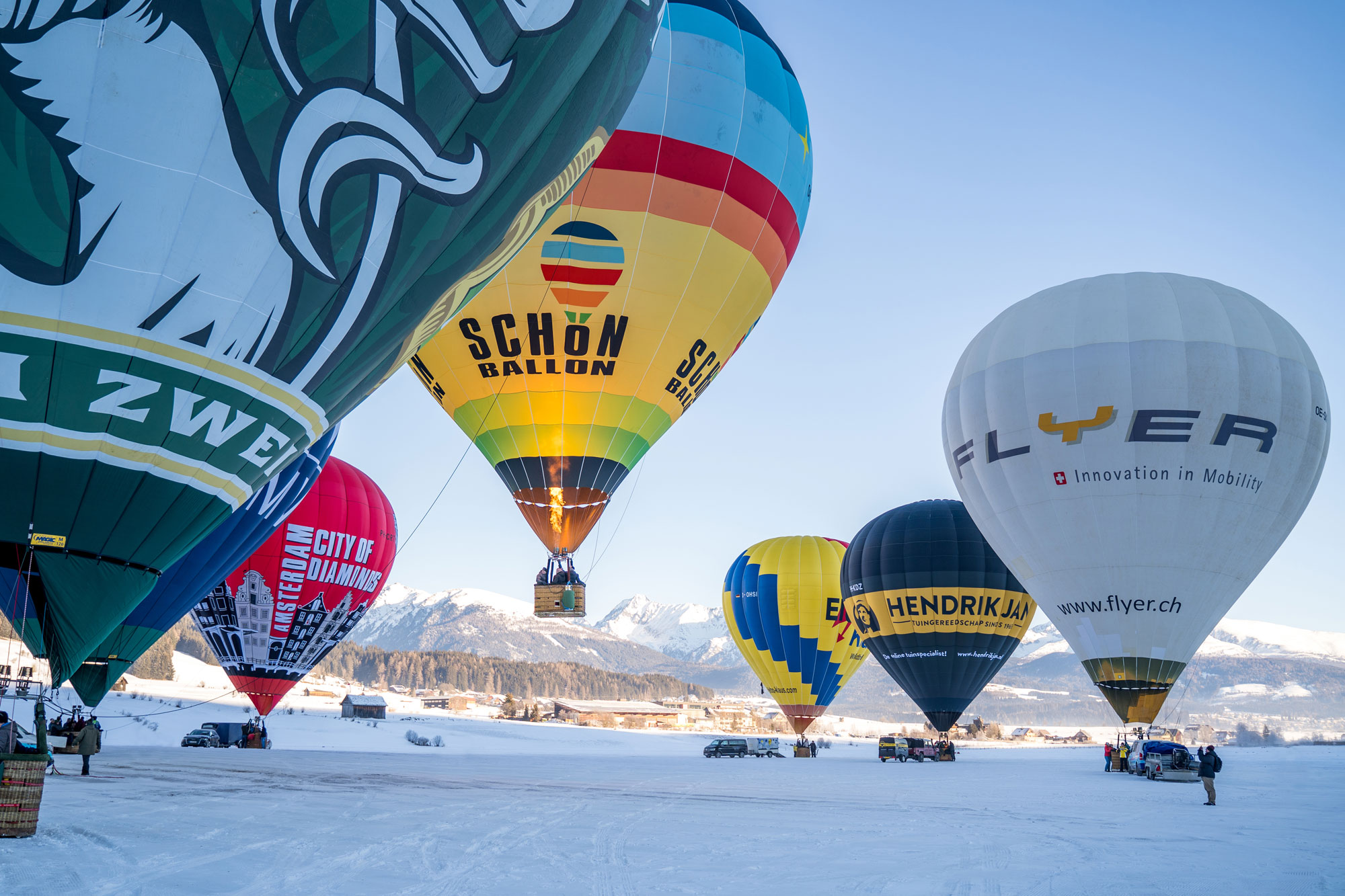Mehrere Heißluftballone auf einem verschneiten Feld in Mauterndorf, ein Ballon hebt ab. Heißluftballonfahrten im Salzburger Lungau als Erlebnis während der Ballonwoche Mauterndorf.