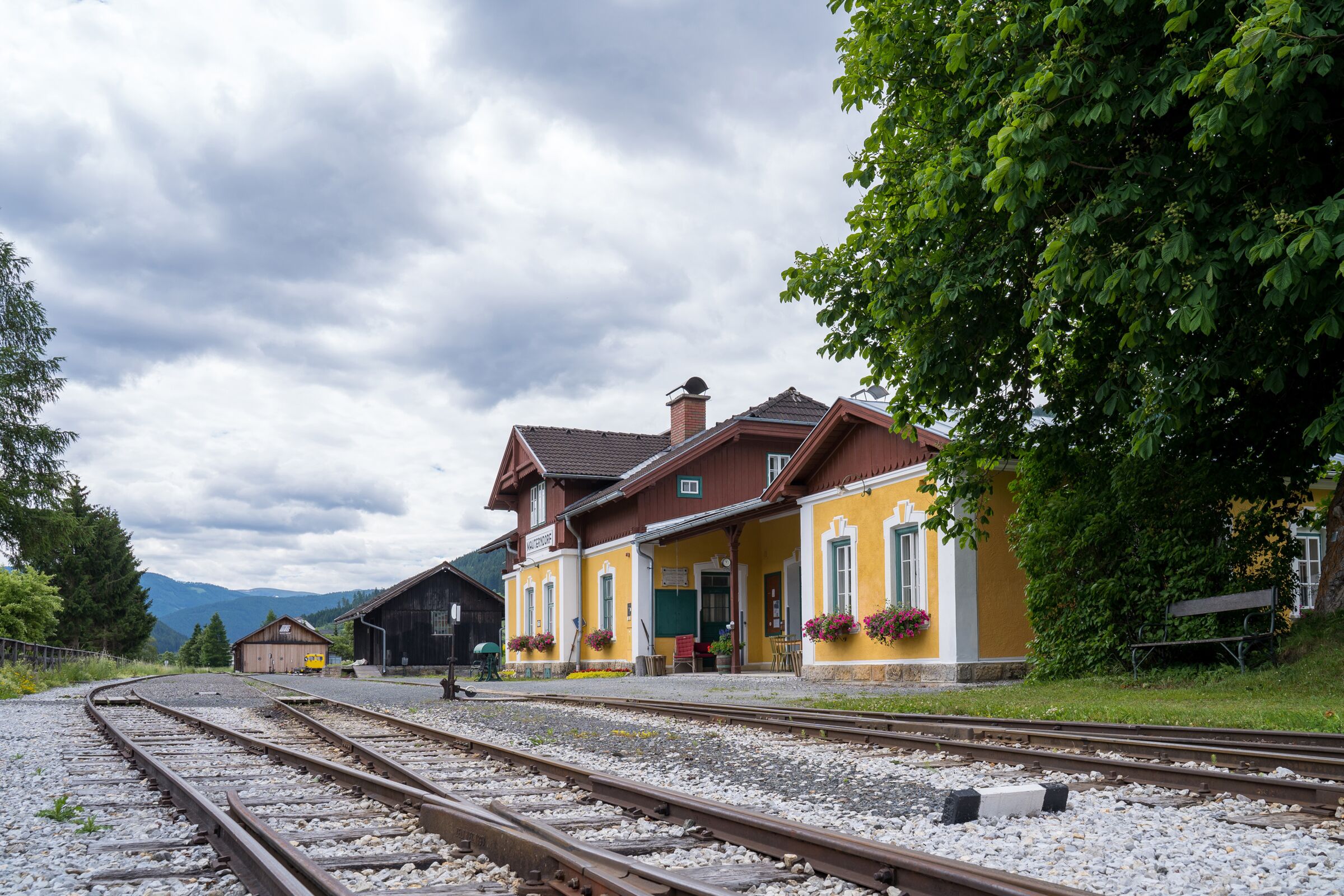 Gelbes Bahnhofsgebäude mit Blumen, umgeben von Natur und Schienen in ländlicher Umgebung.