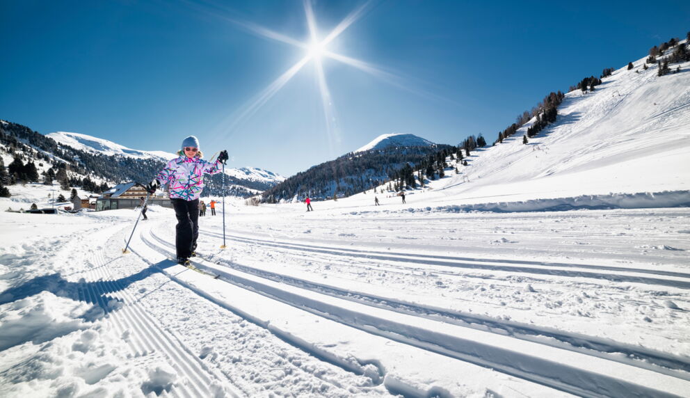 Skifahrer auf sonniger, schneebedeckter Piste vor majestätischen Bergen und blauem Himmel.