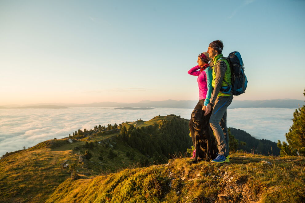 Zwei Wanderer mit Hund genießen den Sonnenaufgang über einer Wolkendecke in den Bergen.