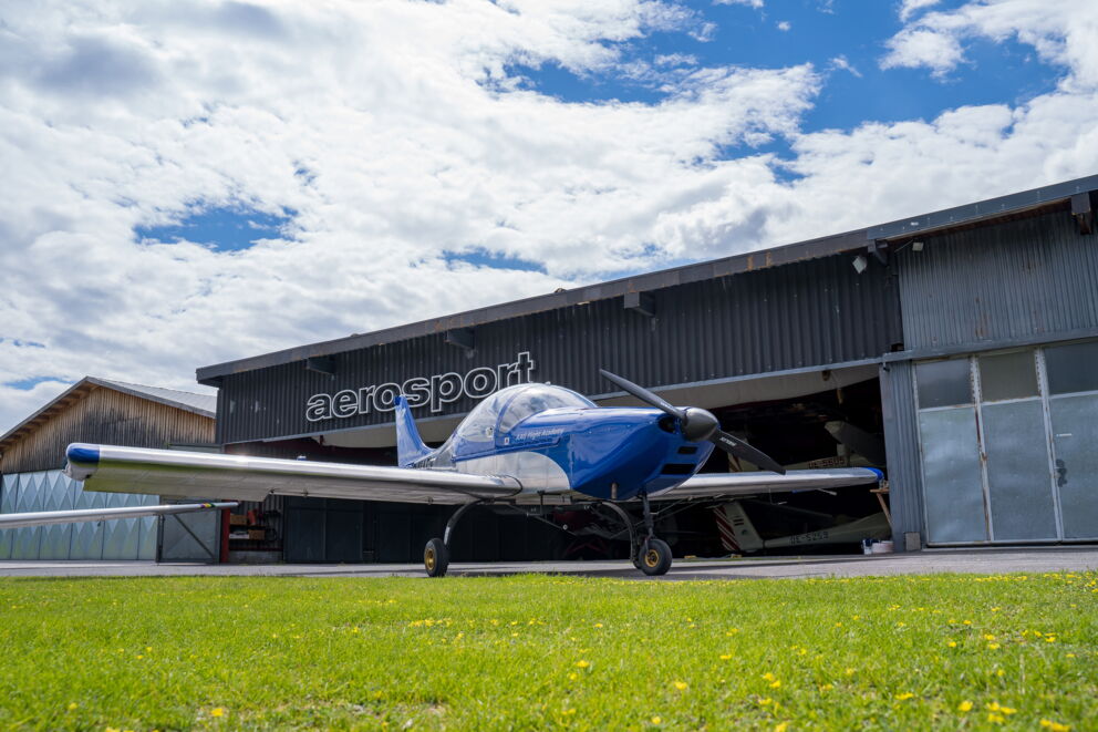 Blauer Sportflieger vor Hangar unter bewölktem Himmel, auf grüner Wiese.