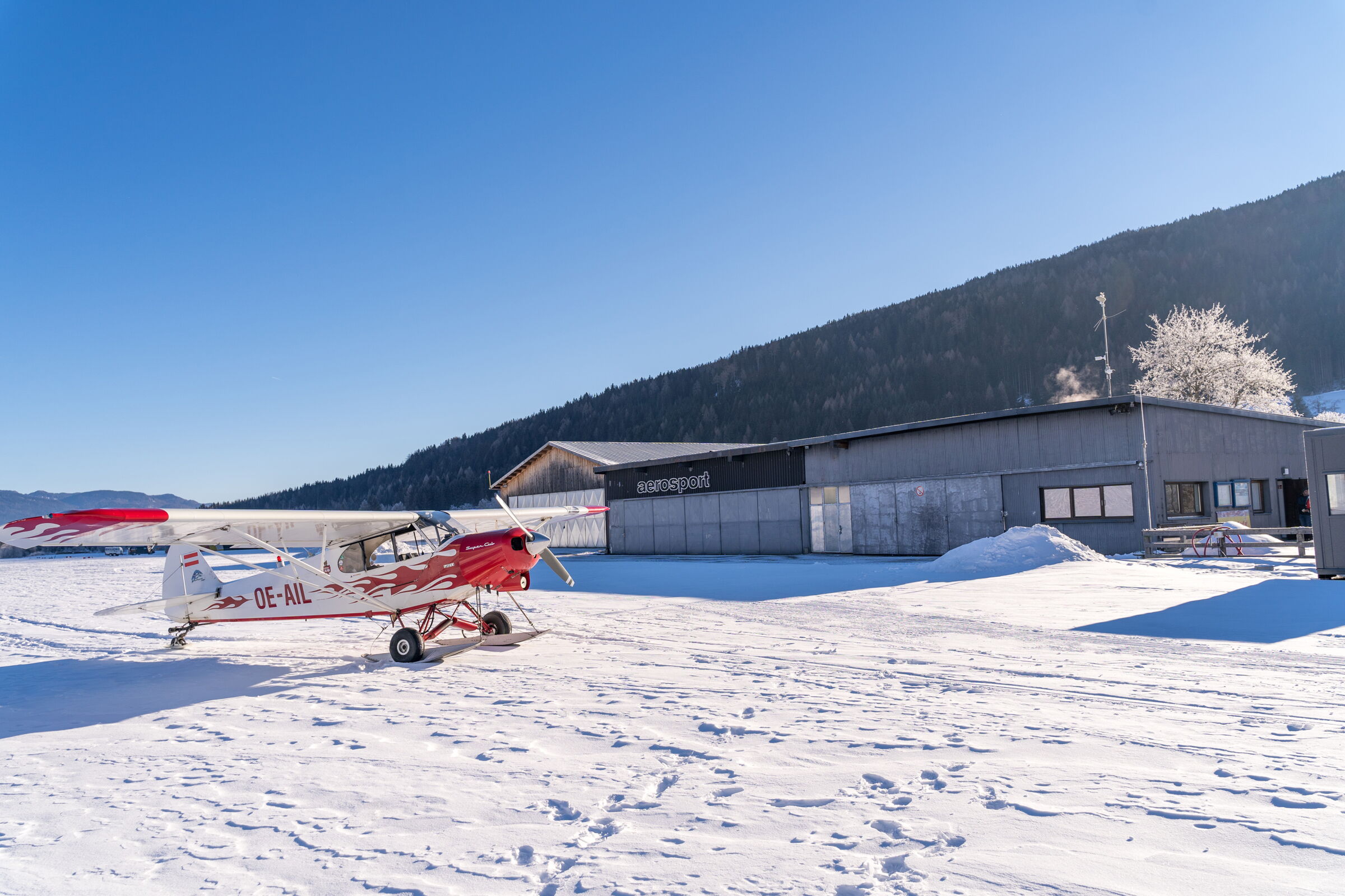 Kleines Flugzeug auf schneebedecktem Feld vor Hangar und bewaldeter Berglandschaft.