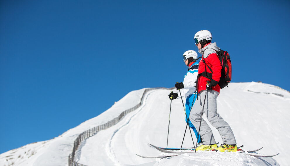 Zwei Skifahrer auf schneebedecktem Hang unter klarem, blauem Himmel.