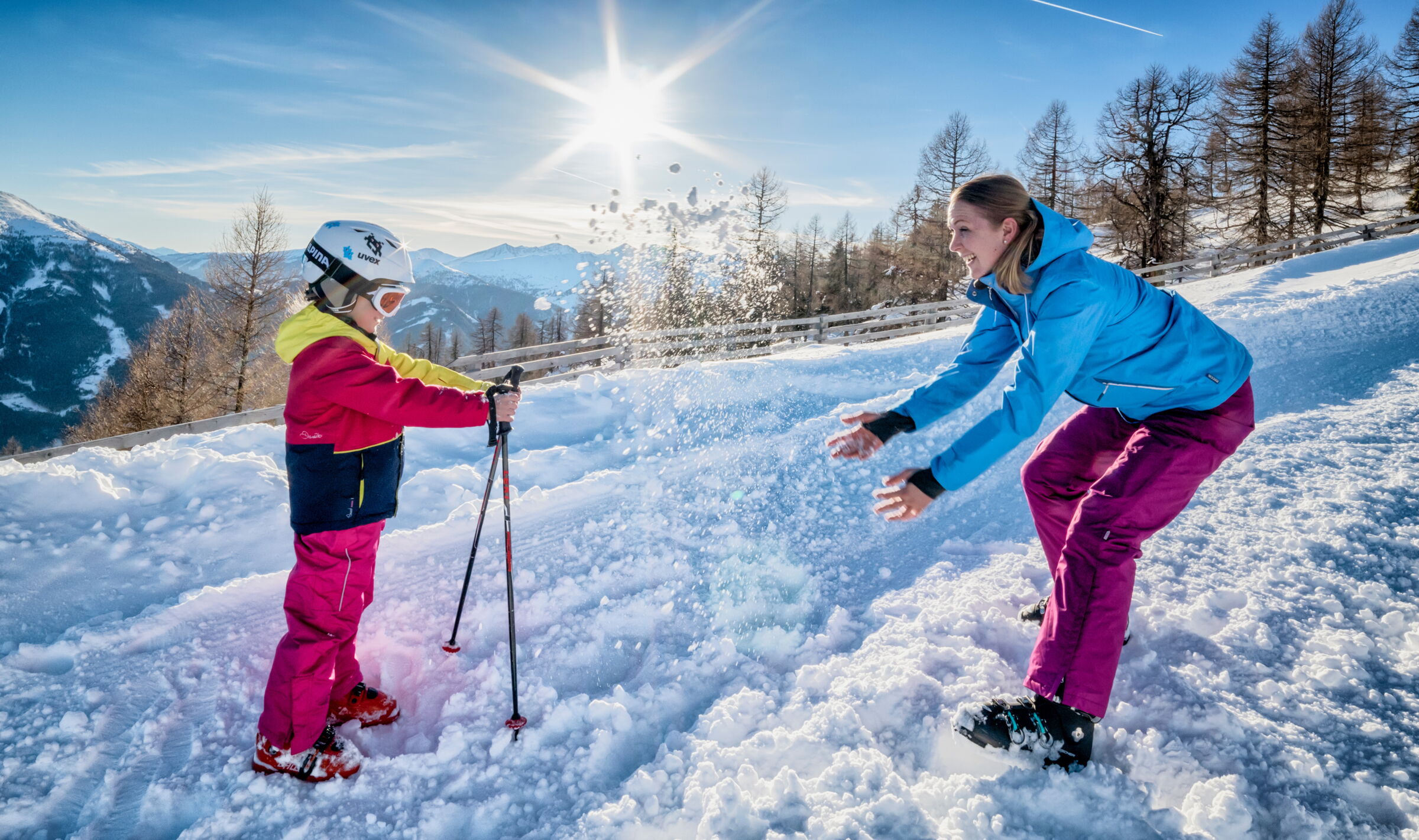 Kind und Erwachsener spielen im Schnee bei strahlendem Sonnenschein in alpiner Landschaft.