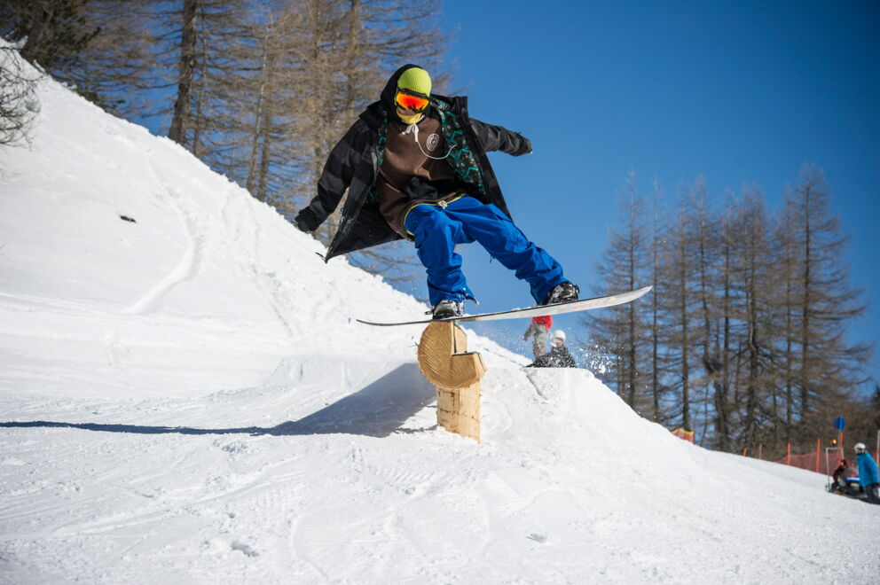 Snowboarder über Holzrampe springend, umgeben von verschneiter Berglandschaft und blauem Himmel.