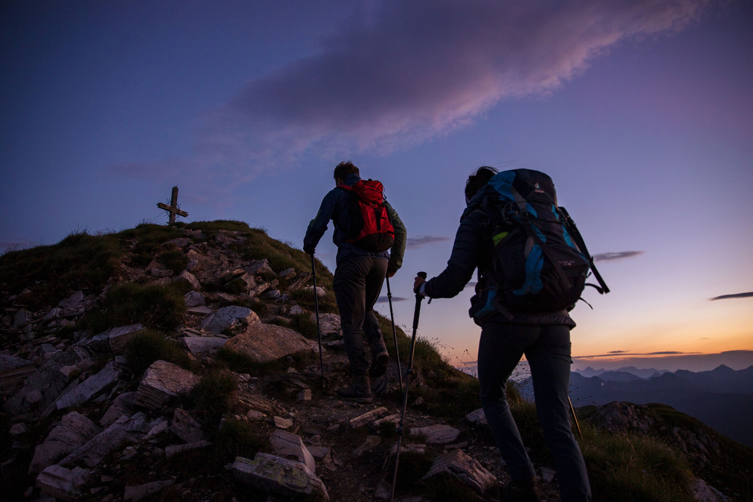 Zwei Wanderer bei Sonnenaufgang auf einem felsigen Bergpfad mit Kreuz.