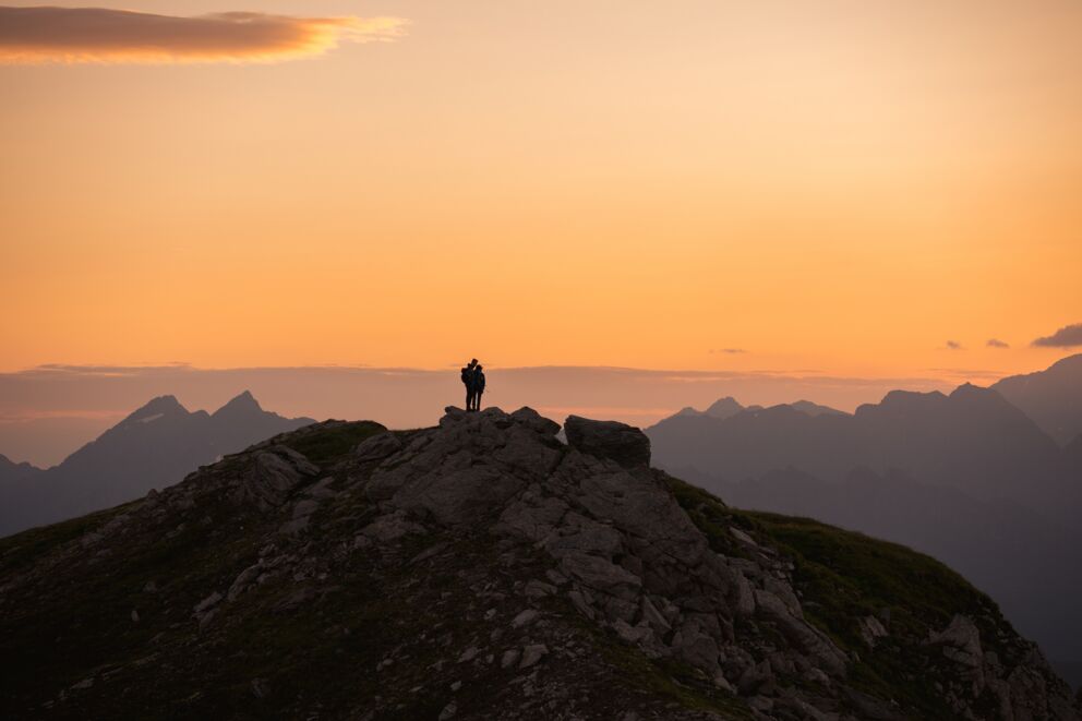 Zwei Wanderer auf Berggipfel bei Sonnenuntergang, mit beeindruckendem Alpenpanorama.