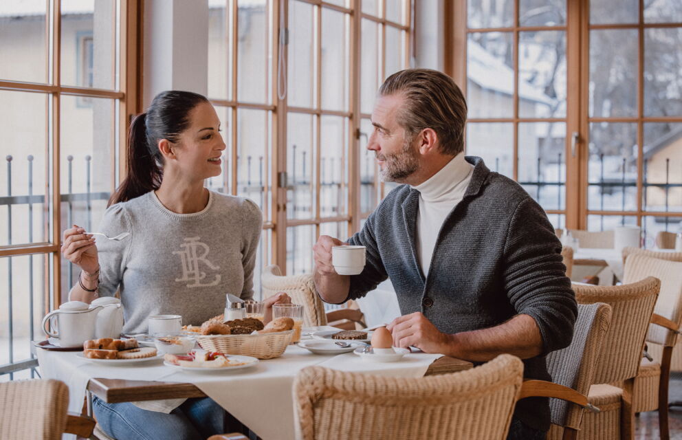 Two people enjoy breakfast in a bright, cozy room with large windows.