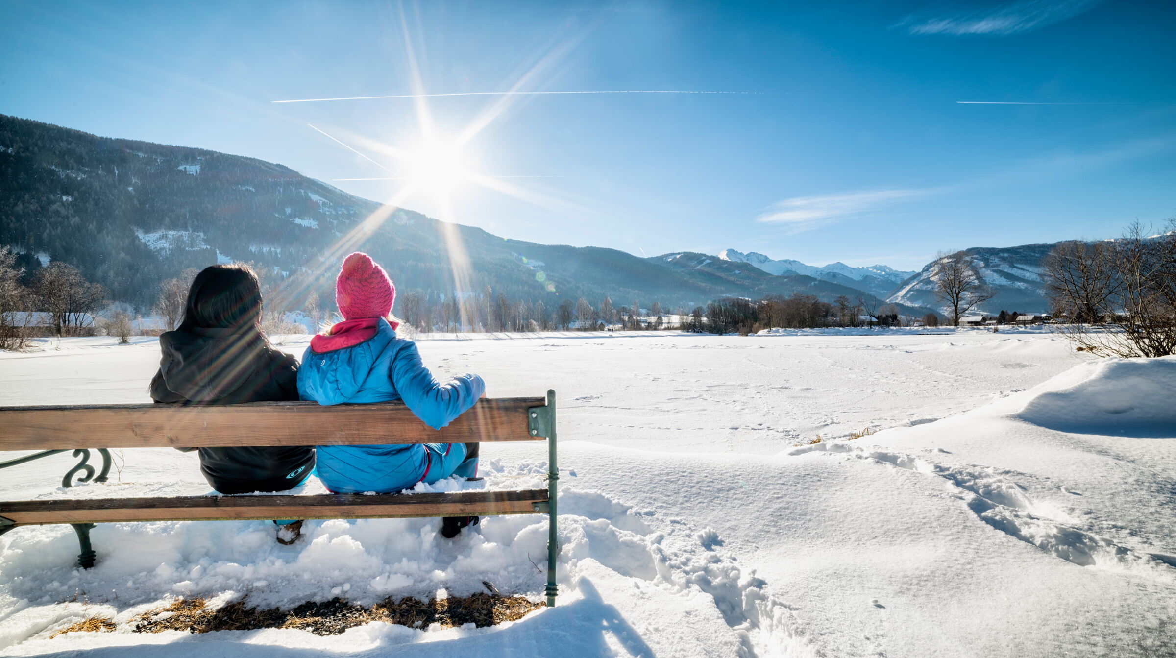 Zwei Menschen auf einer Bank genießen den sonnigen Ausblick auf schneebedeckte Berge.