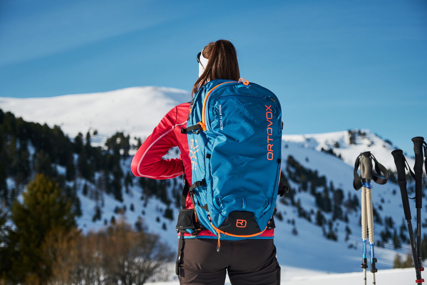 Frau mit blauem Rucksack blickt auf verschneite Berge unter klarem Himmel.