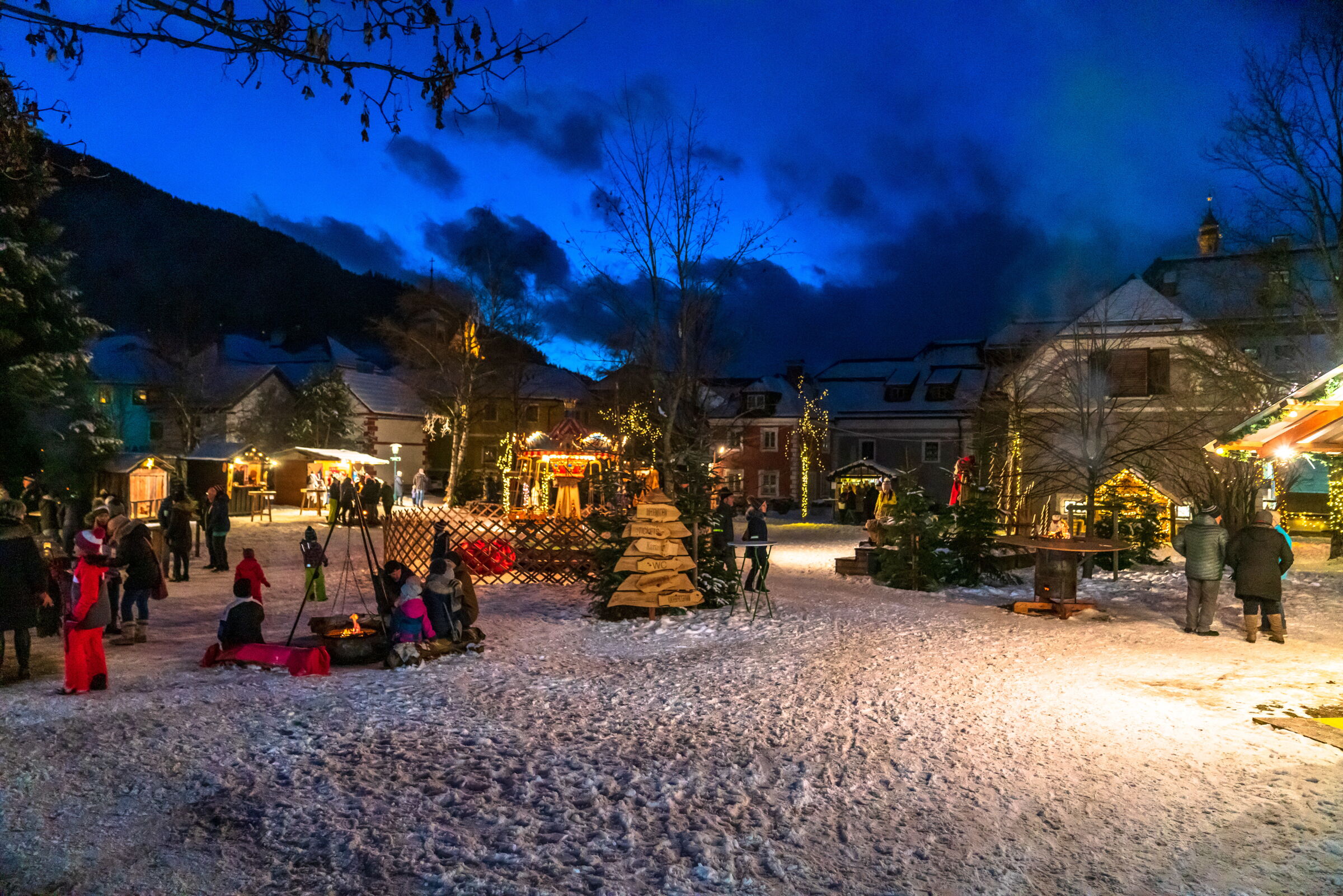 Menschen genießen winterlichen Dorfmarkt mit Lichtern und schneebedecktem Boden.