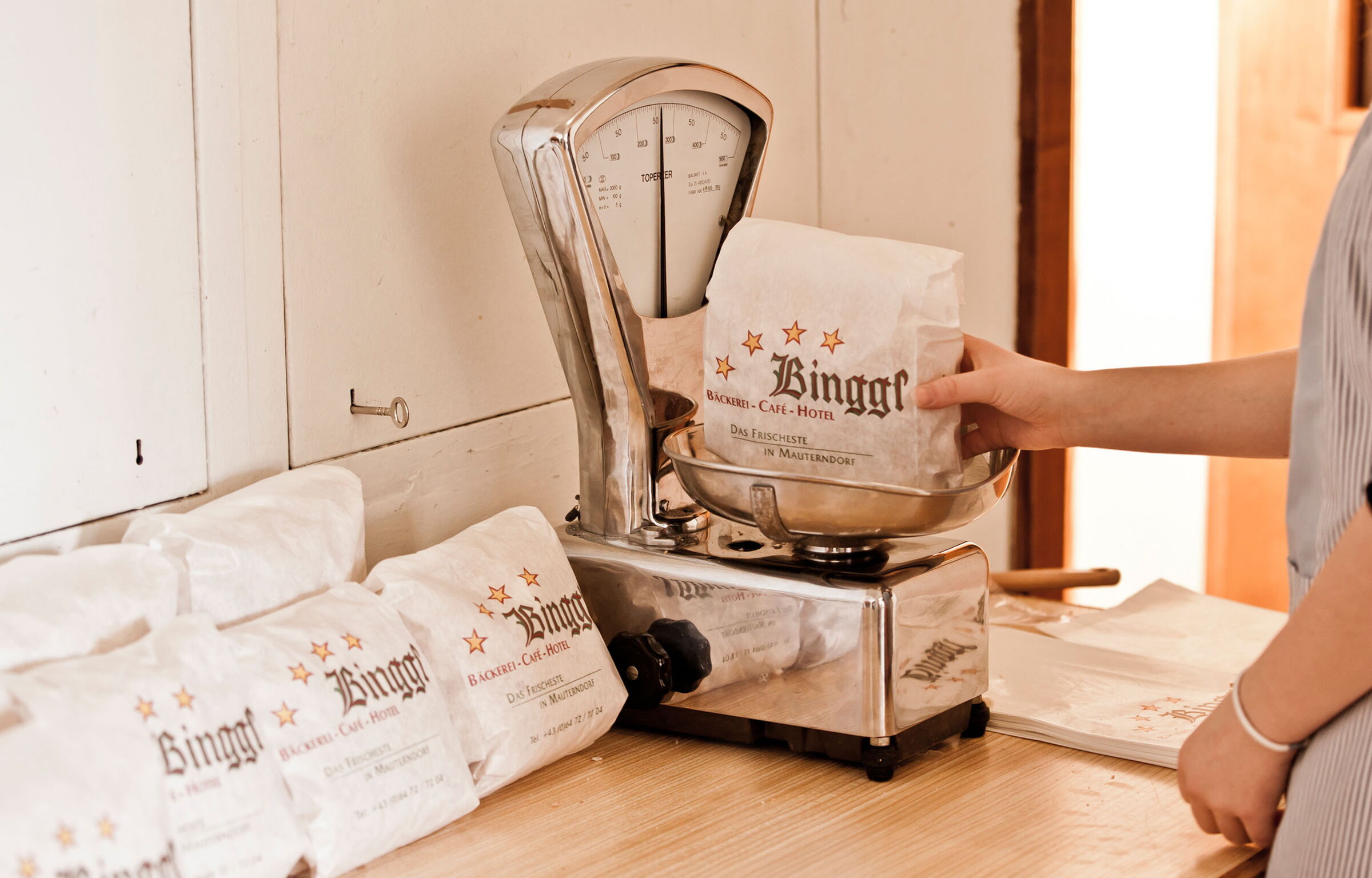 Person wiegt Brot in traditioneller Bäckerei mit alter Waage und Binggl-Verpackung.
