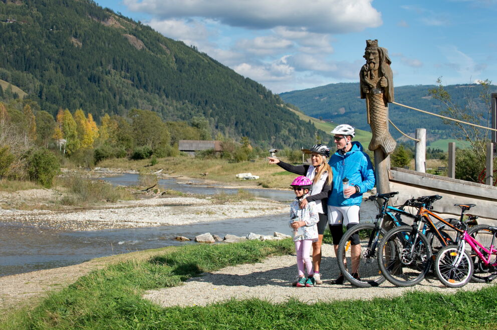 Familie mit Fahrrädern vor malerischer Fluss- und Berglandschaft bei Sonnenschein.