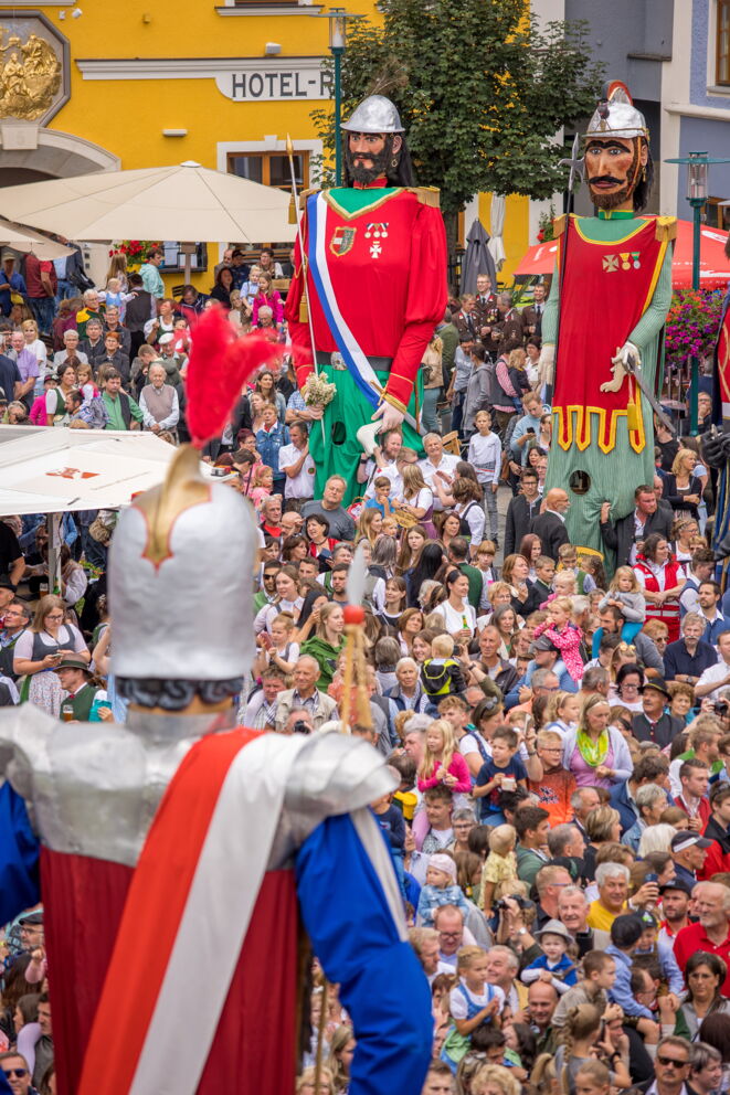 Menschenmenge mit traditionellen Riesenfiguren bei einem bunten Straßenfest.