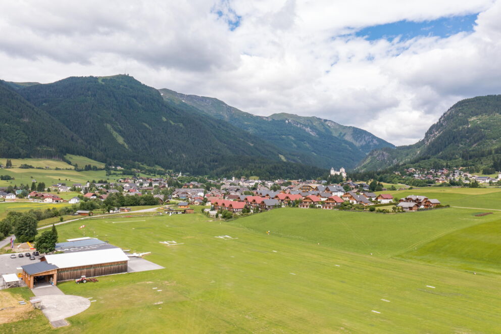 Malerisches Dorf umgeben von grünen Hügeln und majestätischen Bergen unter blauem Himmel.