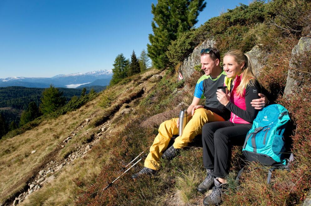 Zwei Wanderer machen Pause mit Blick auf bewaldete Berge und klaren Himmel.