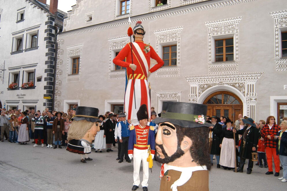 Große Puppenparade in traditioneller Kleidung, umringt von Menschen vor historischen Gebäuden.