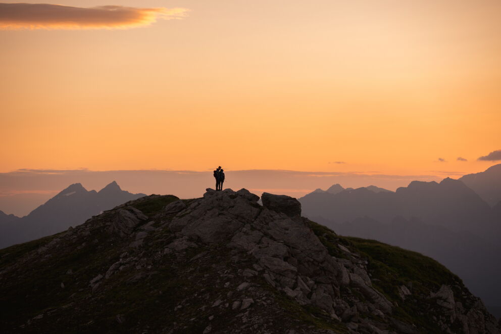 Zwei Wanderer auf Berggipfel bei Sonnenuntergang, mit beeindruckendem Alpenpanorama.