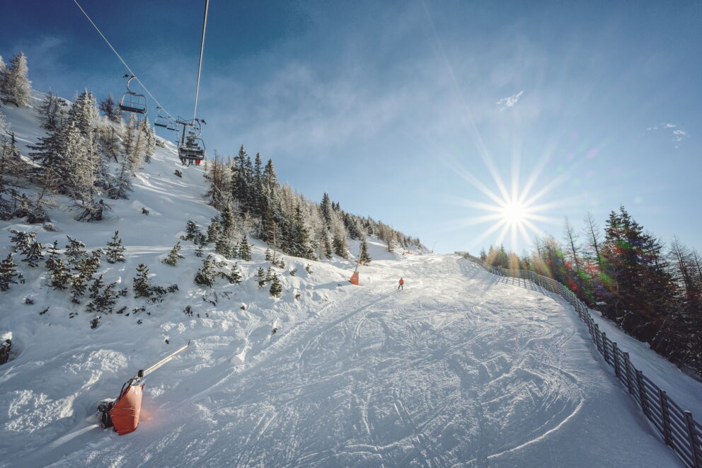 Skifahrer auf sonniger Piste mit Sessellift und verschneiten Bäumen im Hintergrund.