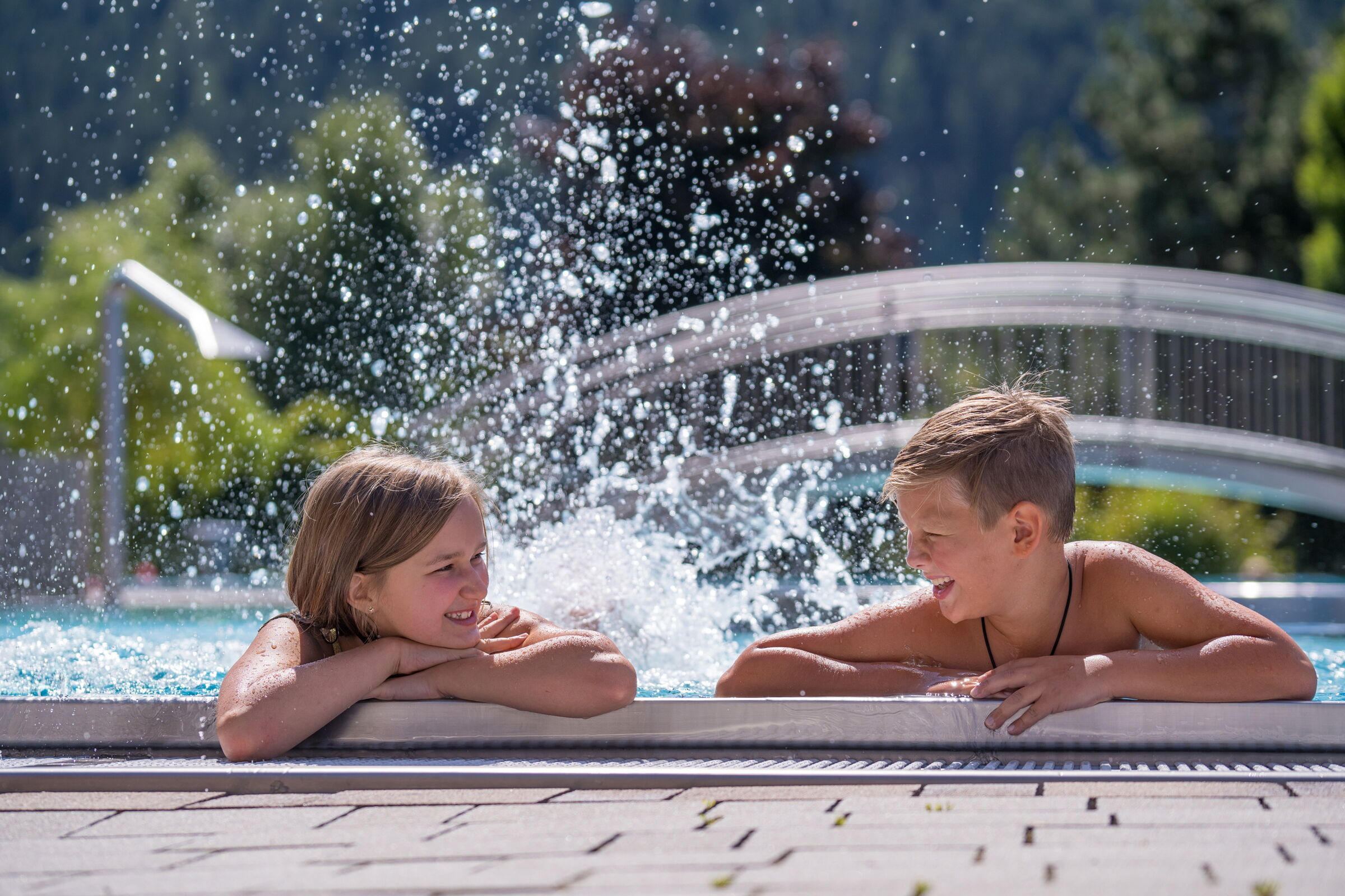 Zwei Kinder genießen den Pool mit spritzendem Wasser im Sonnenschein.