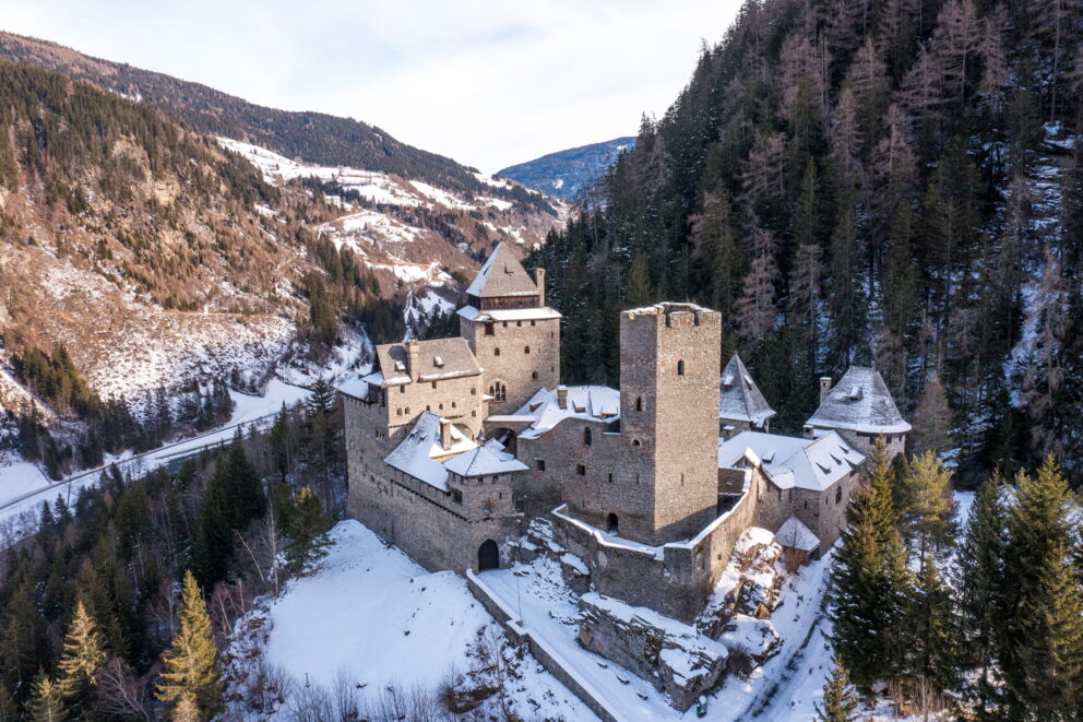 Verschneite Burg inmitten malerischer Berge und dichten Wäldern umgeben von Schnee.
