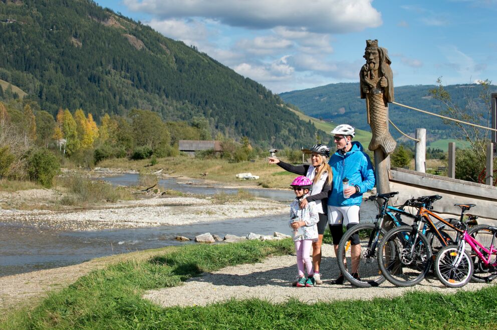 Familie mit Fahrrädern vor malerischer Fluss- und Berglandschaft bei Sonnenschein.