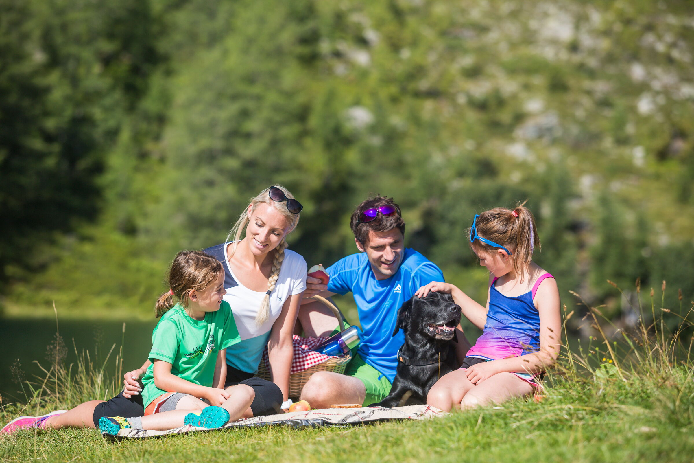 Familie genießt Picknick mit Hund in malerischer Naturkulisse.
