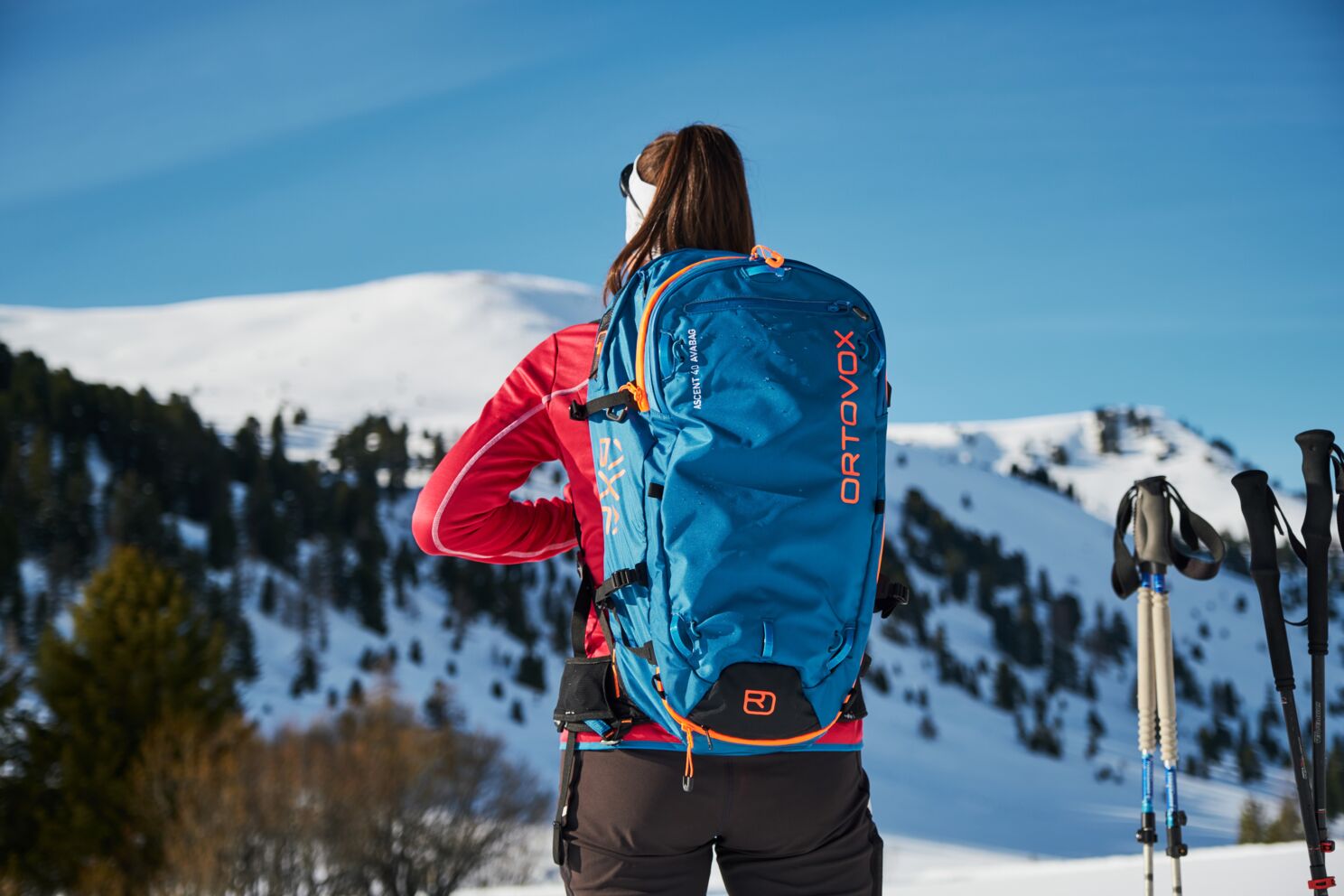 Frau mit blauem Rucksack blickt auf verschneite Berge unter klarem Himmel.