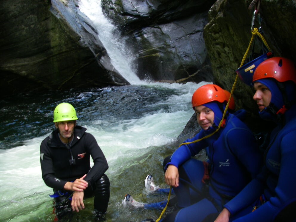 Drei Personen in Neoprenanzügen beim Canyoning vor einem Wasserfall.