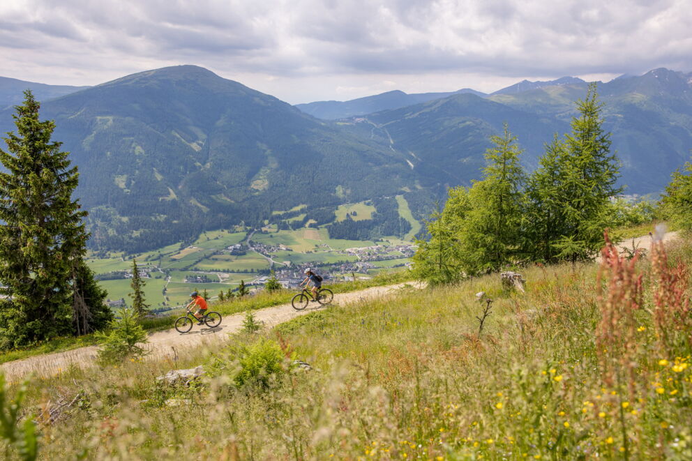 Zwei Mountainbiker auf einem Bergpfad mit weitem Talblick im Hintergrund.