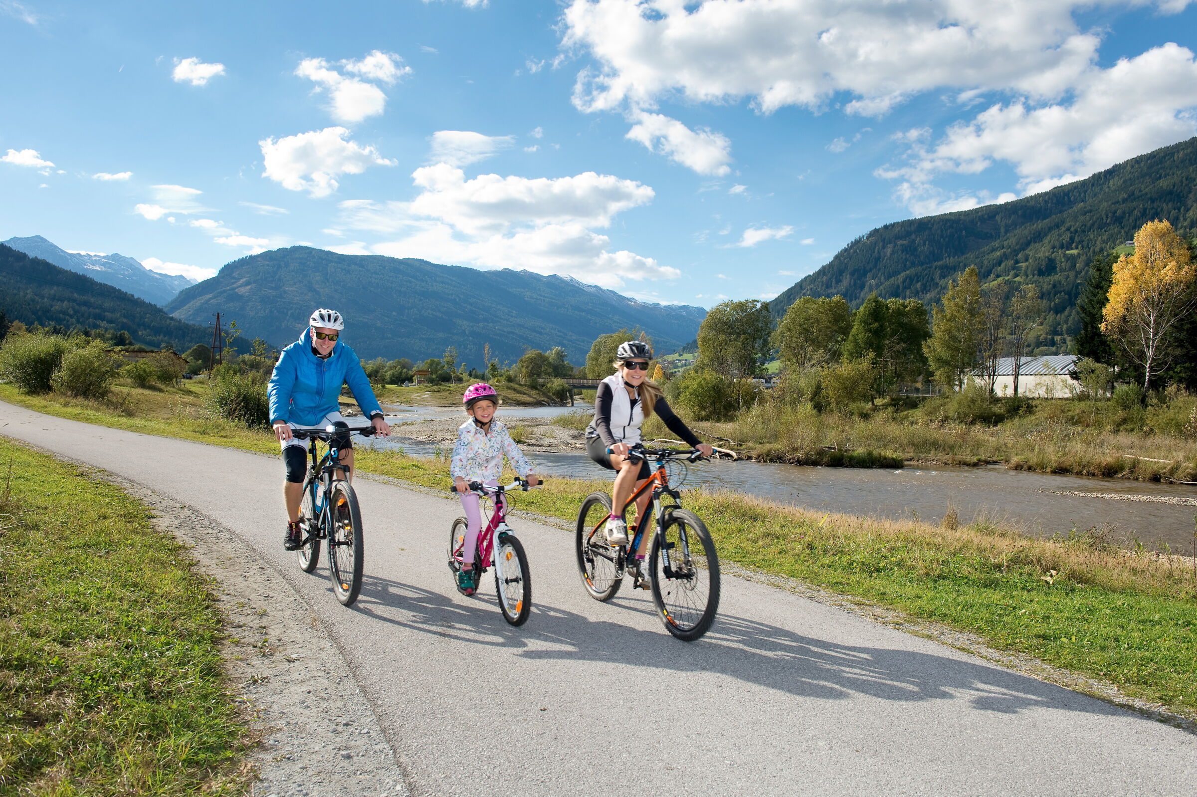 Familie radelt entlang eines Flusses mit beeindruckender Bergkulisse und blauem Himmel.
