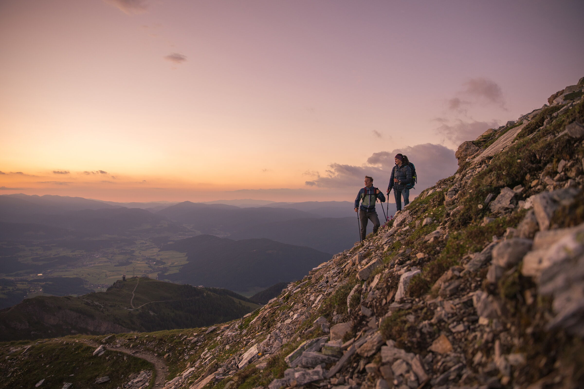Zwei Wanderer steigen bei Sonnenuntergang einen felsigen Berghang hinauf.