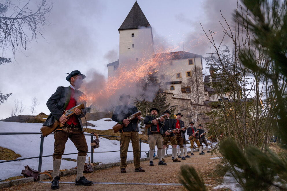 Gruppe traditionell gekleideter Männer zündet Gewehre vor einem historischen Gebäude.