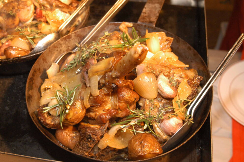 Geschmorte Fleischstücke mit Zwiebeln, Knoblauch und Kräutern in gusseisernen Pfannen serviert.