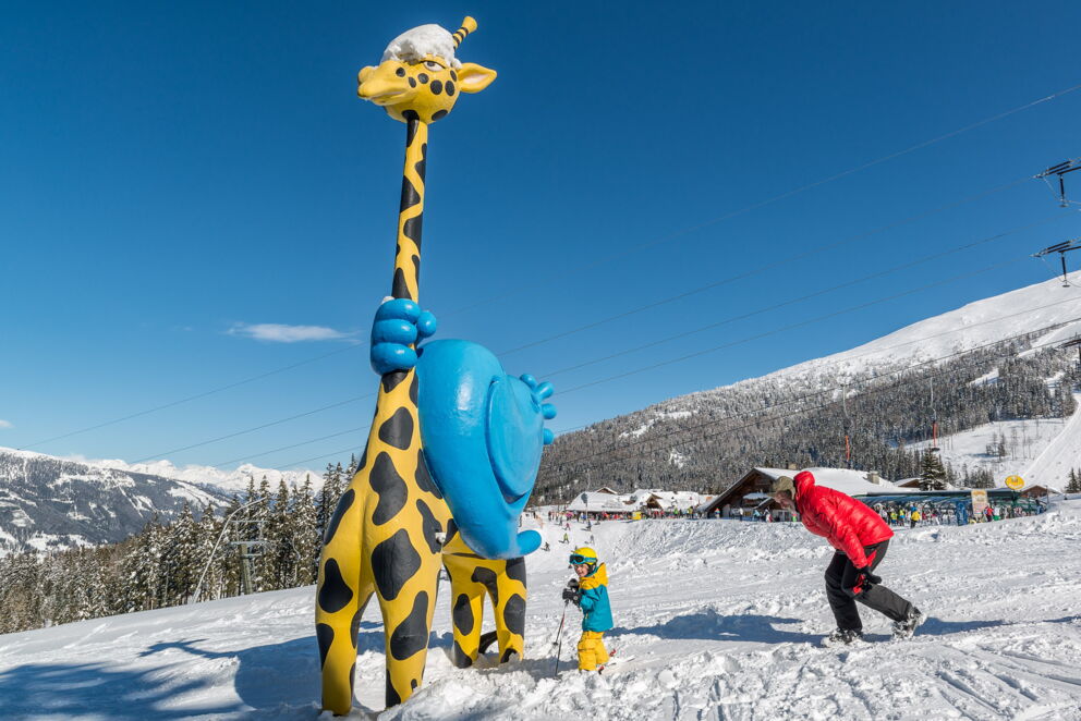 Kind und Erwachsener bei bunter Giraffenfigur im Schnee unter blauem Himmel