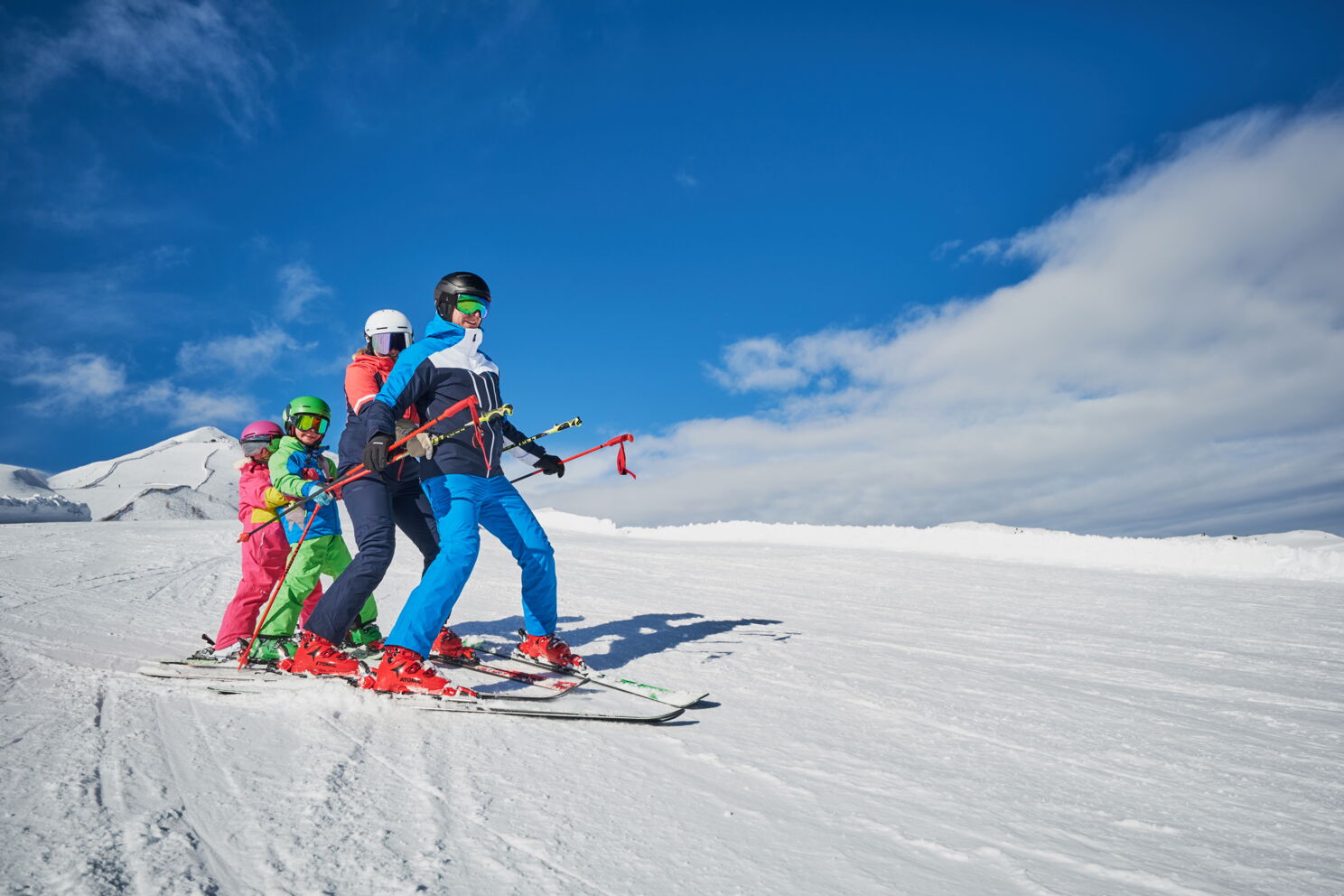 Familie genießt gemeinsames Skierlebnis auf sonniger, schneebedeckter Piste unter blauem Himmel.
