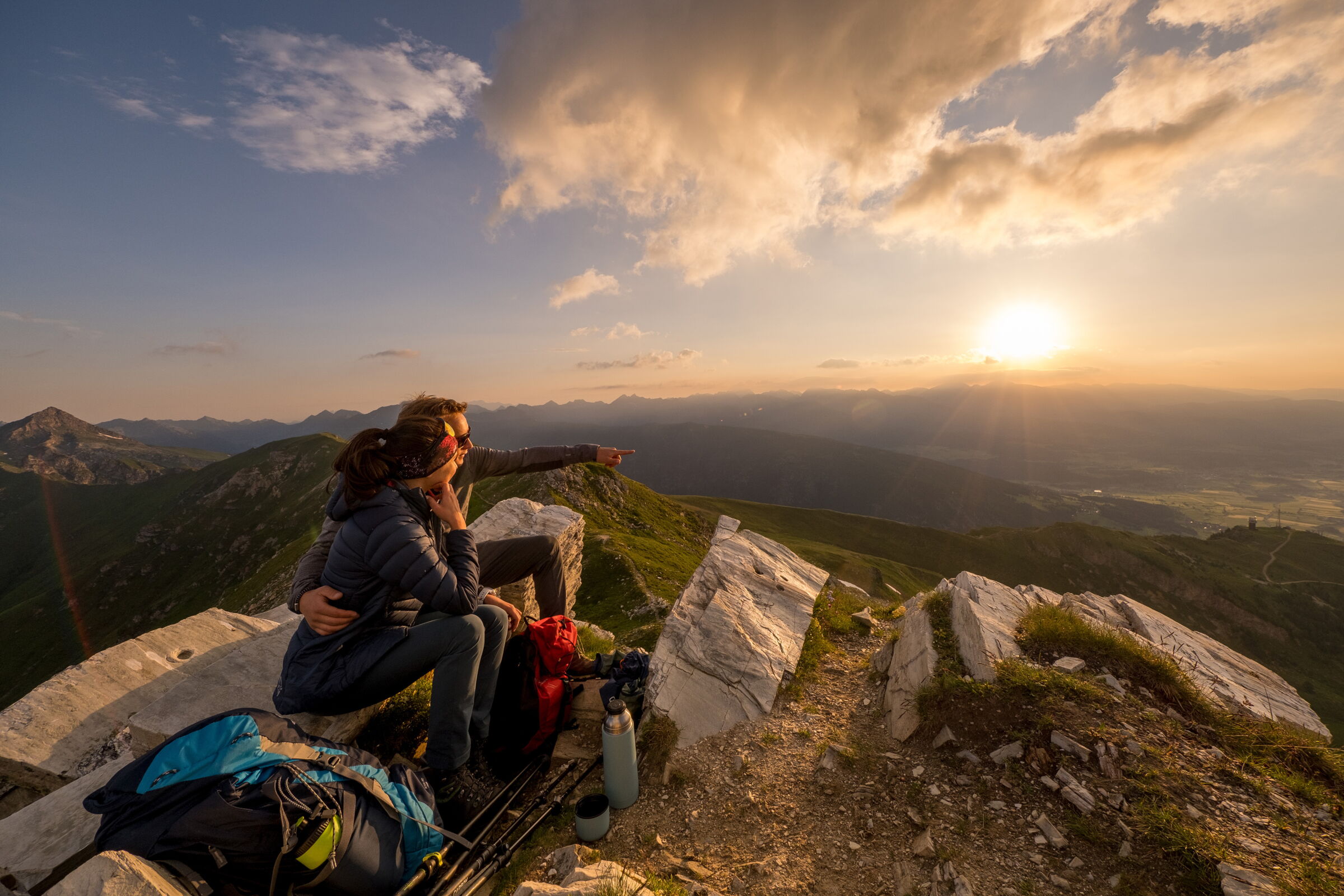 Zwei Personen genießen den Sonnenuntergang auf einem Berggipfel mit weitem Talblick.