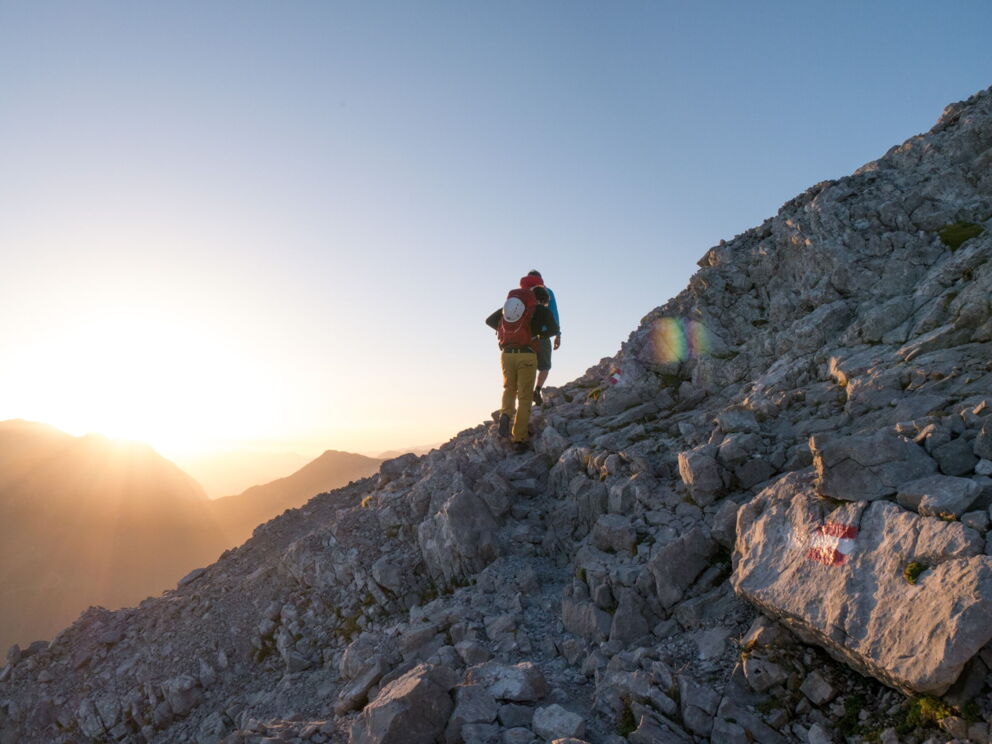 Zwei Wanderer auf steinigem Pfad bei Sonnenaufgang, umgeben von Bergen.