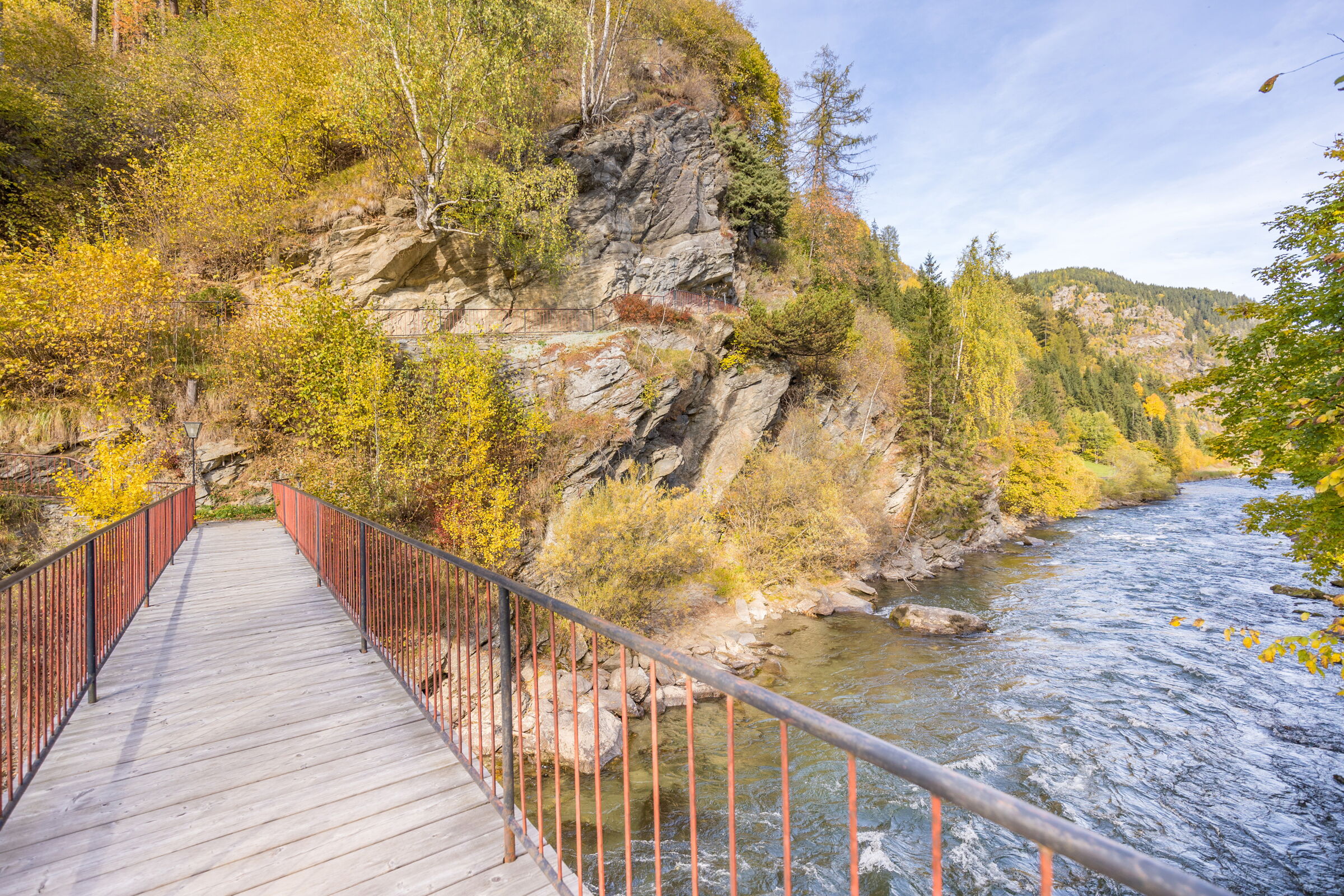 Holzbrücke über einen Fluss, umgeben von herbstlichen Bäumen und Felsen.