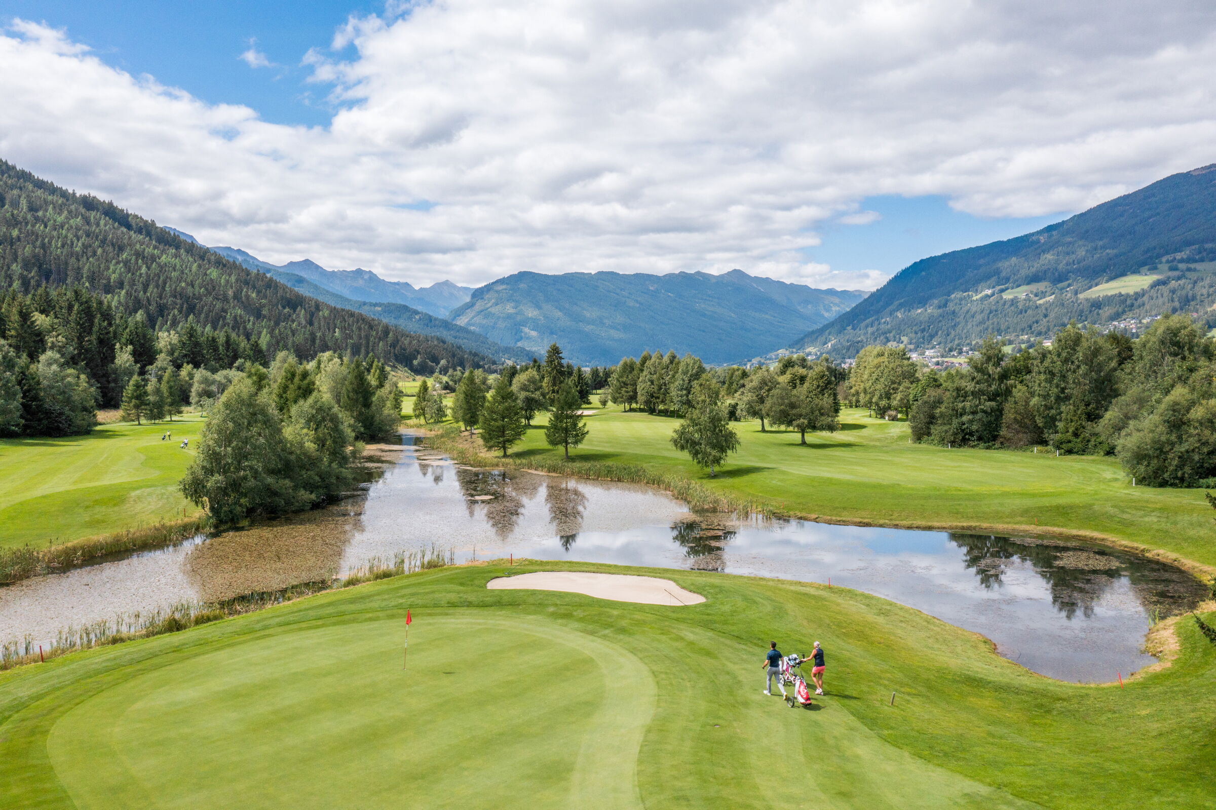 Golfplatz mit Teich, umgeben von Bergen und Wäldern unter blauem Himmel.