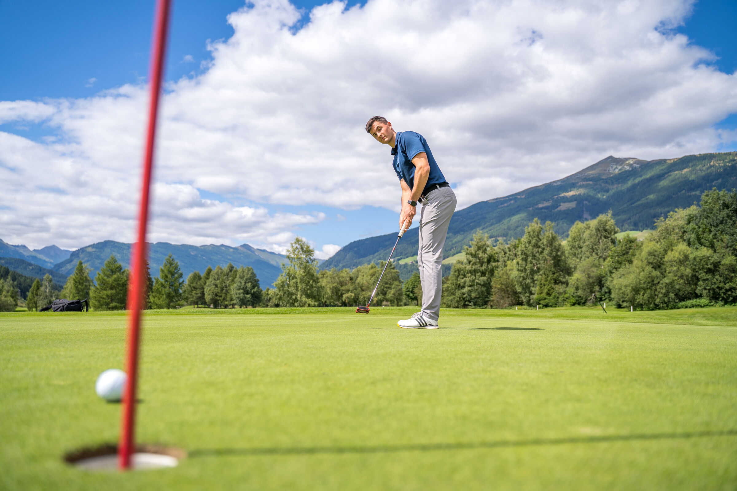 Golfer auf gepflegtem Grün mit majestätischer Berglandschaft im Hintergrund unter blauem Himmel.