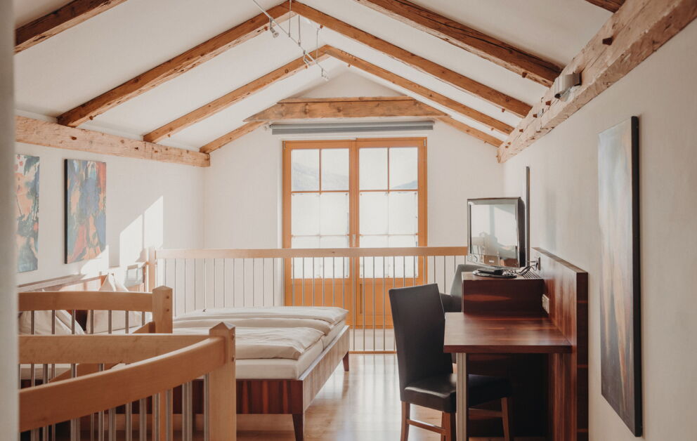 Helles Dachzimmer mit Holzbalken, Schreibtisch und großem Fenster für natürlichen Lichteinfall.