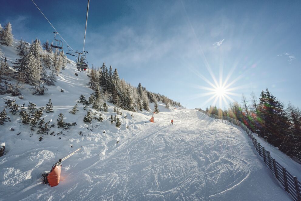 Skifahrer auf sonniger Piste mit Sessellift und verschneiten Bäumen im Hintergrund.
