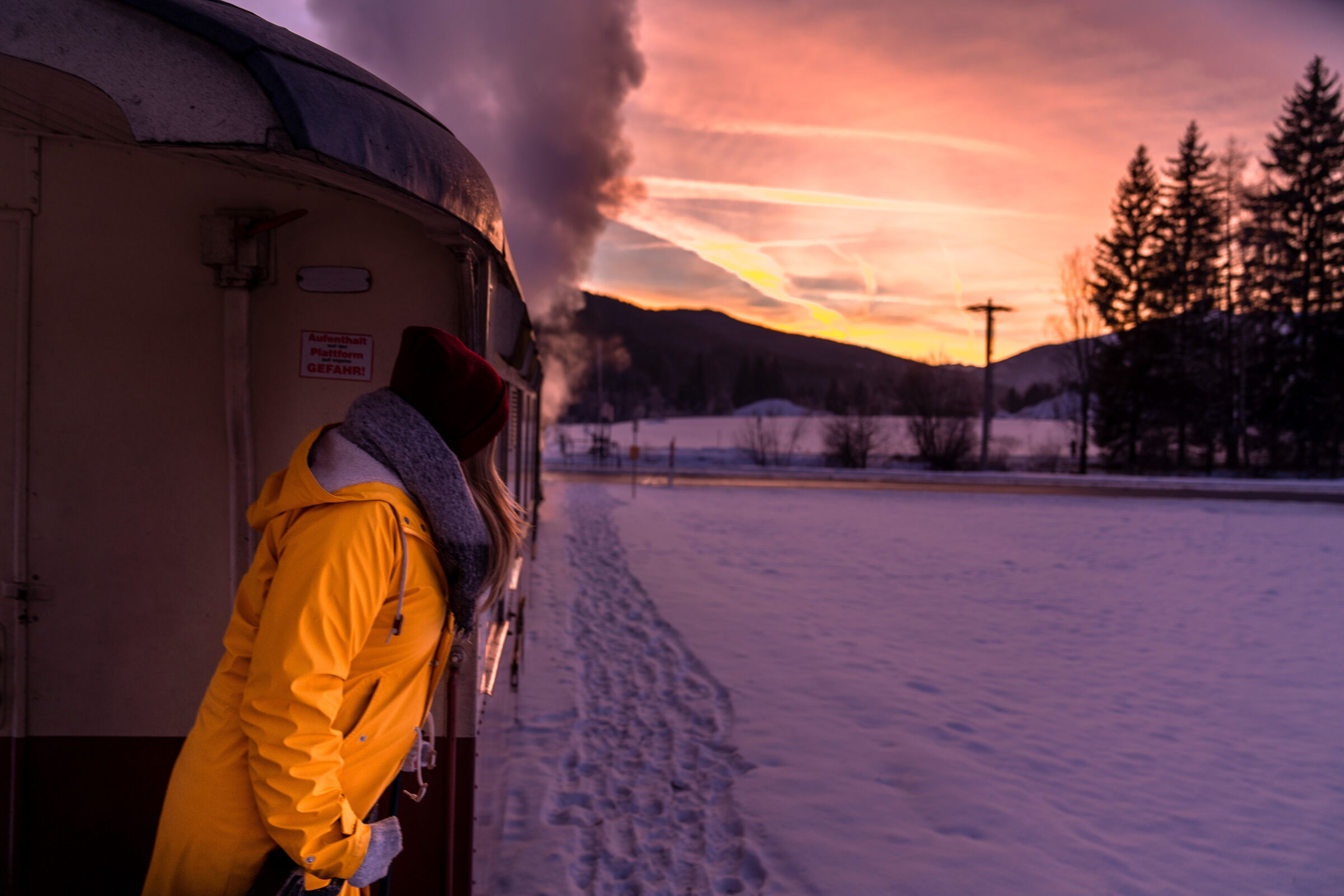 Person im gelben Mantel schaut aus Zug beim Sonnenuntergang auf verschneite Landschaft.
