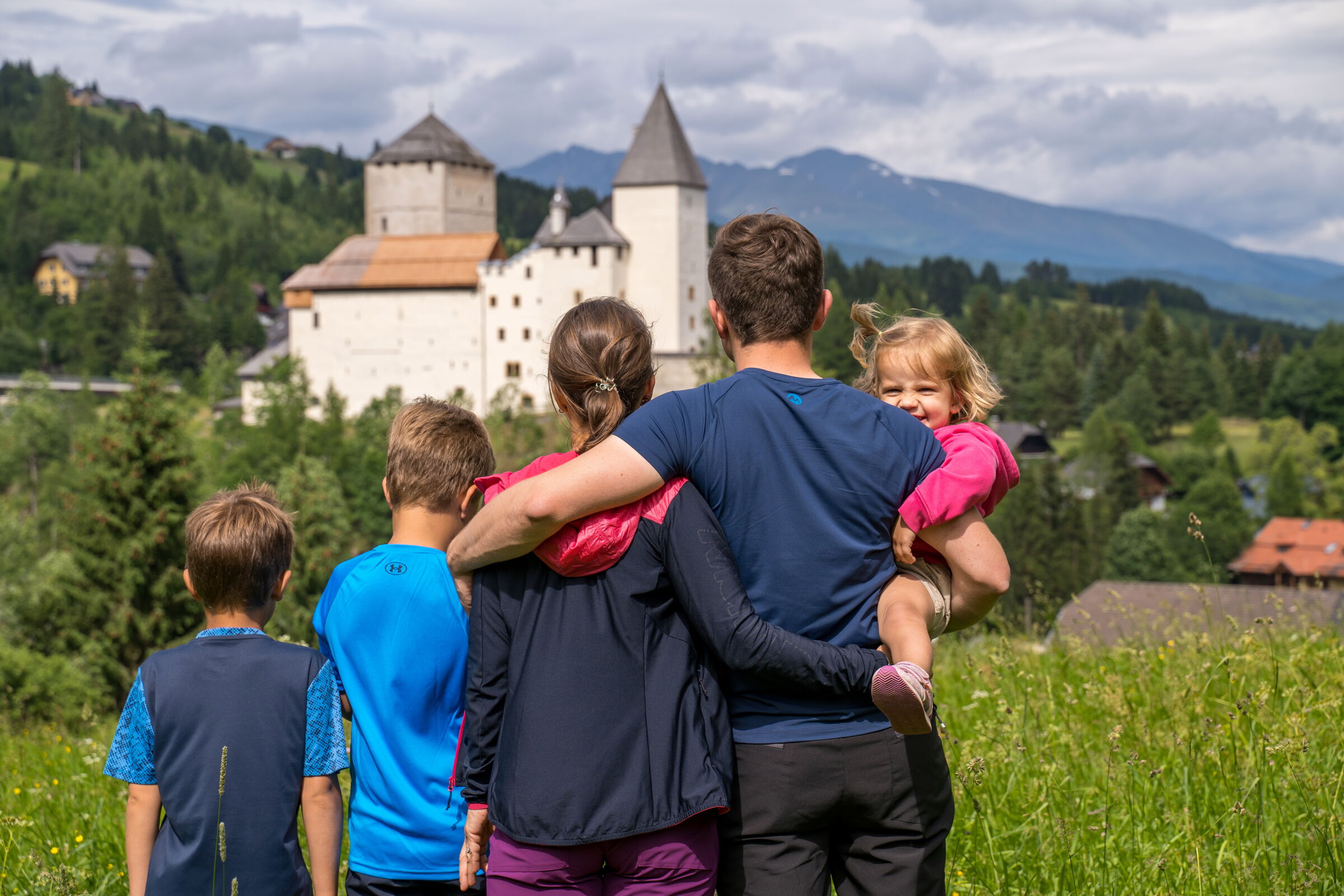 Familie schaut auf eine historische Burg inmitten grüner Landschaft und Berge.
