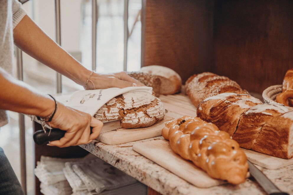 Person schneidet frisches Brot auf Holzbrett in einer Bäckerei.