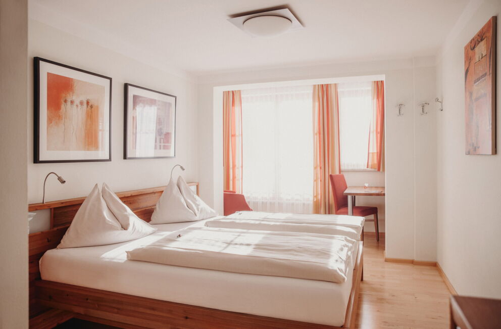 Helles Zimmer mit Holzmöbeln, Bett und gemütlichem Sitzbereich vor Fenstern.