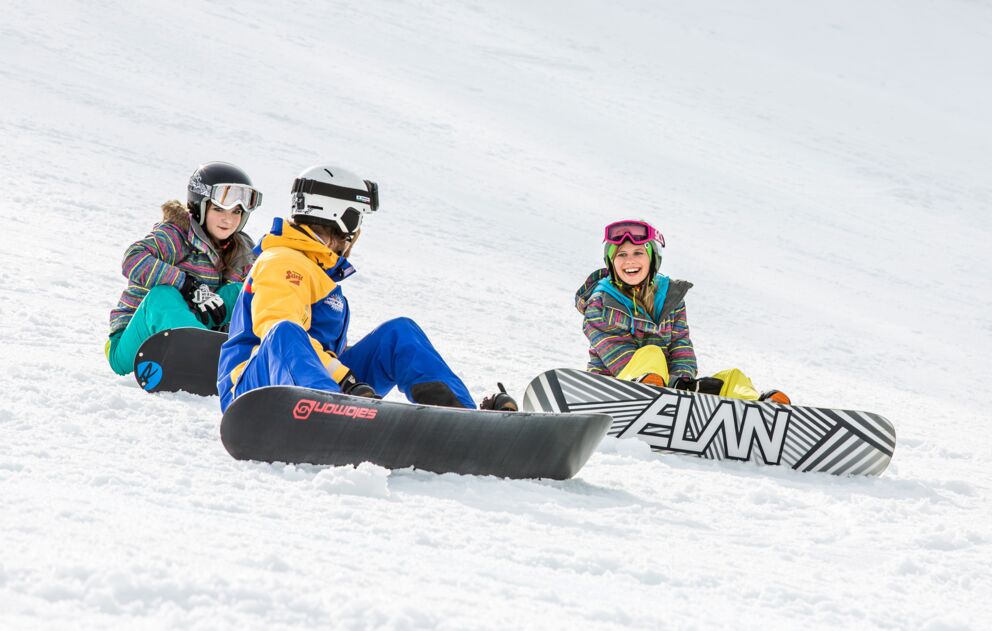 Drei lachende Personen sitzen mit Snowboards im glitzernden Schnee.