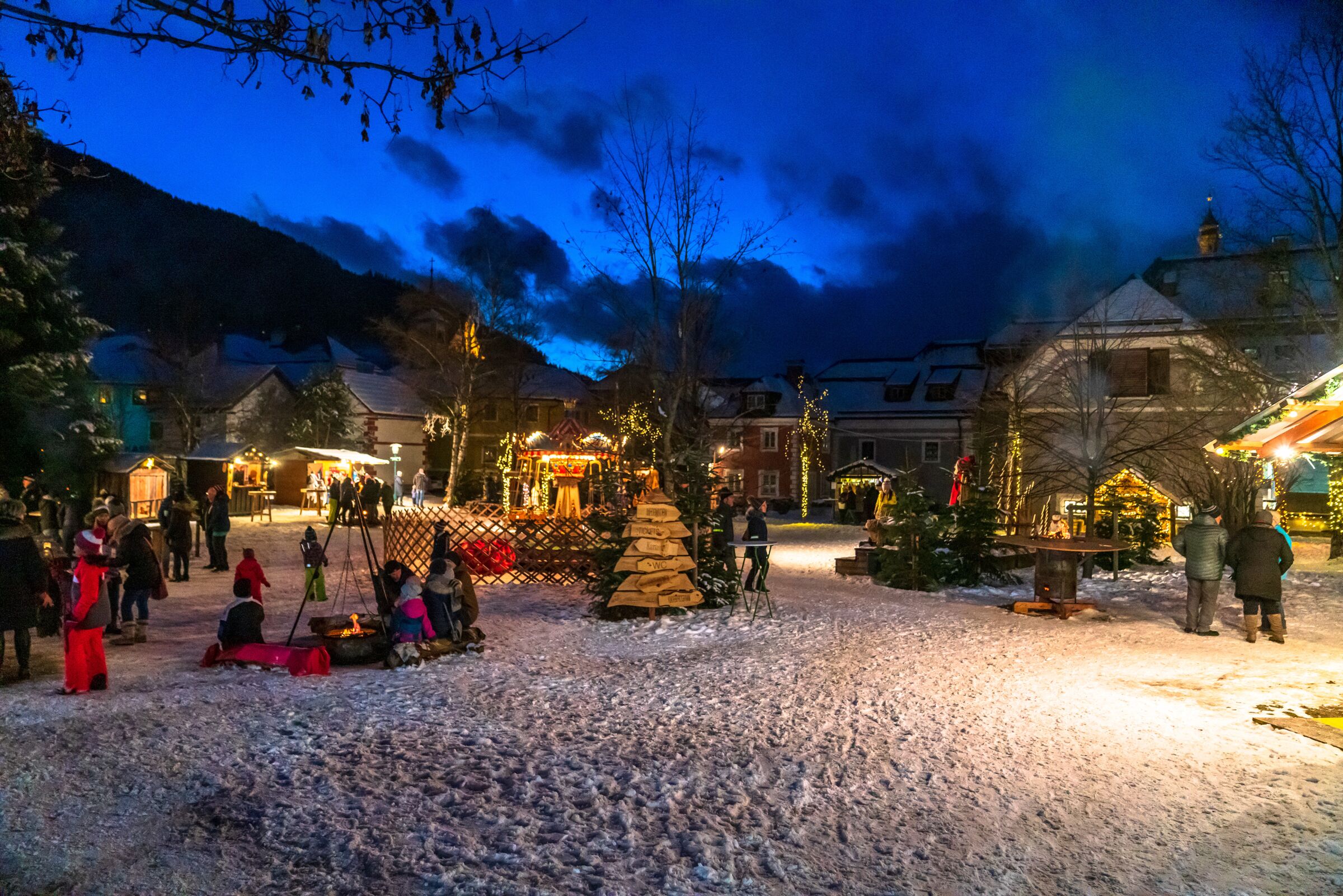 Menschen genießen winterlichen Dorfmarkt mit Lichtern und schneebedecktem Boden.