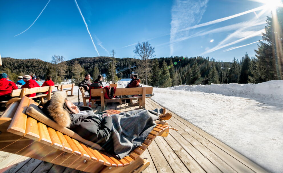 Menschen entspannen auf einer Holzterrasse in winterlicher Berglandschaft unter klarem Himmel.